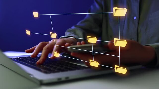 Person works on laptop while holding smartphone with folder icons shown on screen.