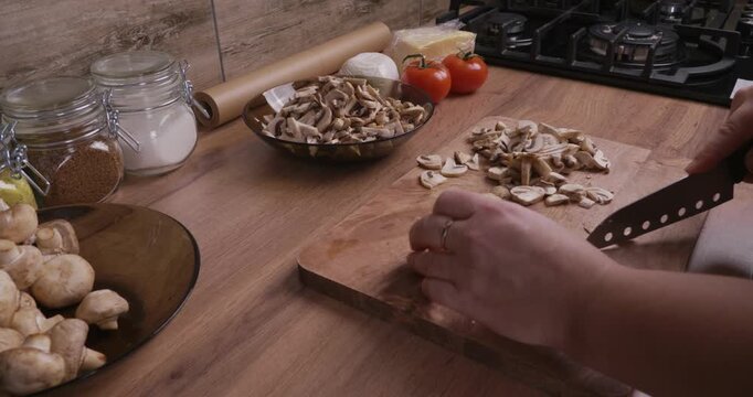 Female chef preparing a healthy meal, slicing fresh champignon mushrooms with a sharp knife on a wooden cutting board in a cozy home kitchen, with other fresh ingredients nearby.