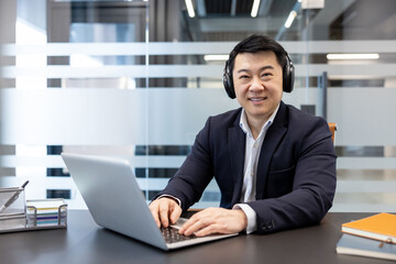 Fototapeta premium Asian businessman smiling while wearing headphones and typing on a laptop, working at a modern office desk with a transparent partition in the background
