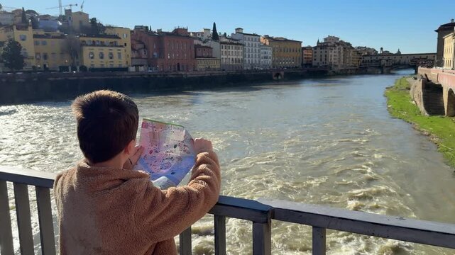 Child looks at map in Florence while facing the river and city buildings under blue sky