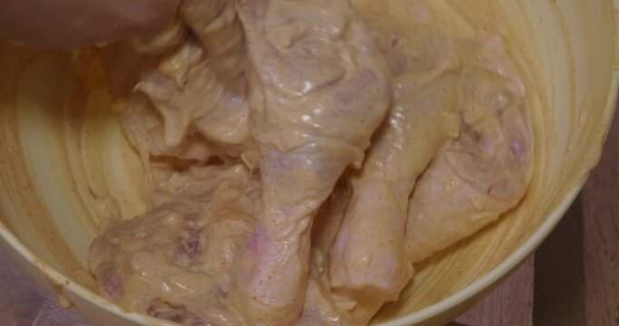 Close-up view of a woman's hands in disposable gloves marinating raw chicken drumsticks in a bowl with a spicy yogurt-based sauce, preparing a delicious and flavorful meal at home.