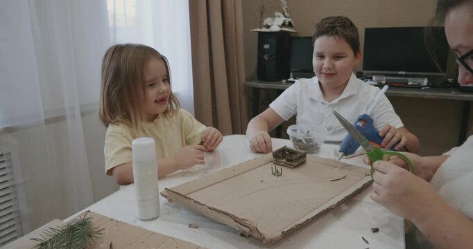 Creative mother with her son and daughter making a craft project from natural materials at a table, using a glue gun and scissors to build a miniature model on a cardboard base.