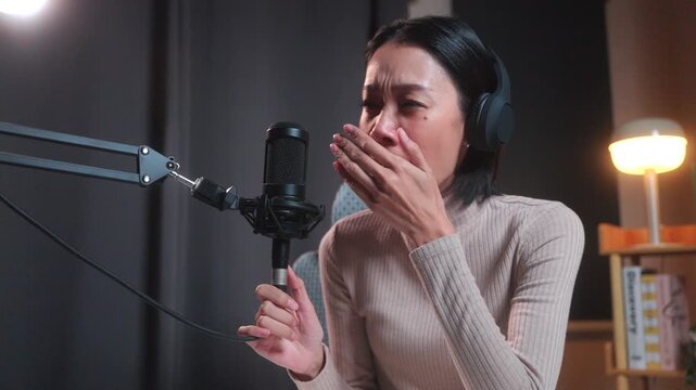 Smiling beautiful asian female podcaster in headphones holding a digital tablet and stylus at her desk in a modern home studio, preparing for a live podcast recording.