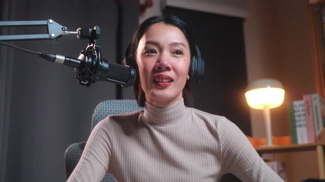 Smiling beautiful asian female podcaster in headphones holding a digital tablet and stylus at her desk in a modern home studio, preparing for a live podcast recording.