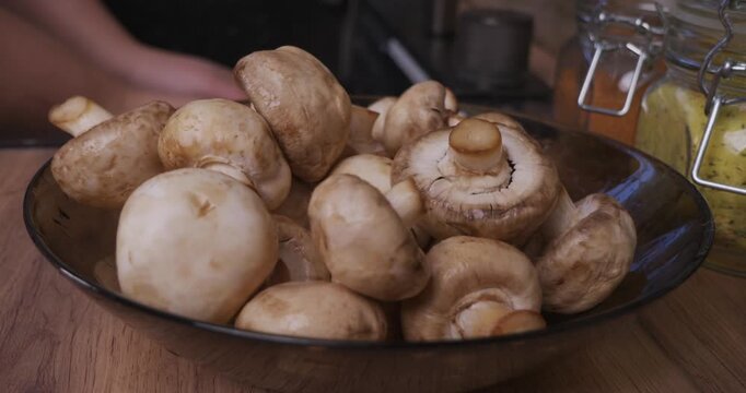 Woman carefully cleans fresh champignon mushrooms in a glass bowl on a wooden kitchen counter, preparing the raw ingredients for cooking a healthy and delicious homemade meal.