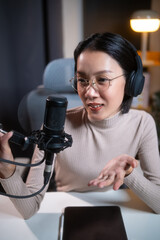 Fototapeta premium Professional beautiful asian female content creator in glasses and headphones recording a podcast at her desk, interacting with a microphone in a modern, well-lit home studio setup. Vertical Shot