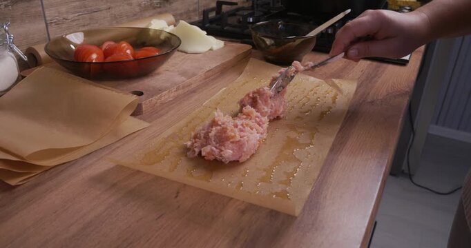 A woman's hands spoon seasoned ground meat onto oiled parchment paper. A woman prepares ingredients for a homemade dinner at a wooden kitchen counter, arranging fresh vegetables.
