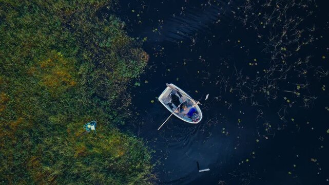 Top-down aerial view of a man and his dog launching a white rowboat from a marshy shoreline and beginning to row across a calm farm pond using wooden oars.