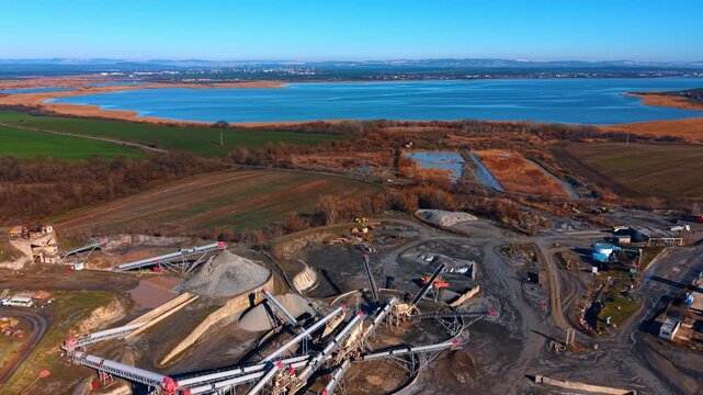 Wide aerial shot of aggregate processing plant and stockpiles. Complex network of conveyor belts and large piles of crushed stone at a quarry site with distant lake.