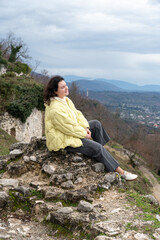 Fototapeta premium Woman sitting on stone ruins overlooking New Athos valley from Anacopia fortress in Abkhazia under cloudy sky, Travel lifestyle scene suitable for tourism marketing, outdoor destination promotion