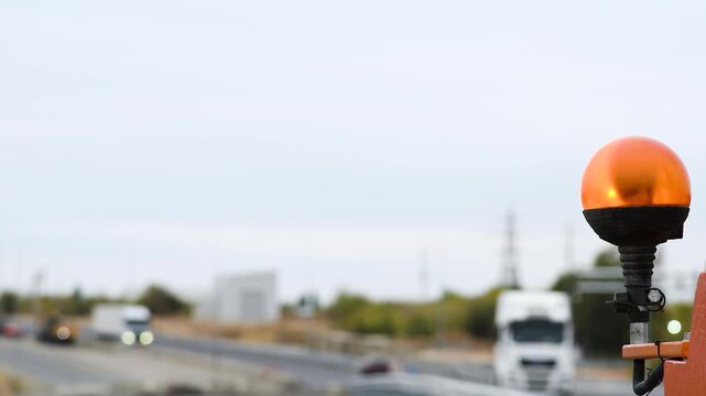 A bright orange warning light mounted on construction equipment, against the background of a highway and cars moving along it.