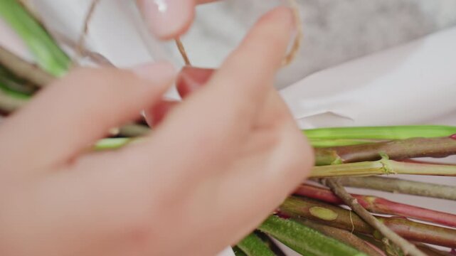 Florist hands tying bouquet stems with twine, closeup showing delicate fingers wrapping paper around green stems, rustic jute knot detail, artisanal workshop atmosphere and careful finishing touch
