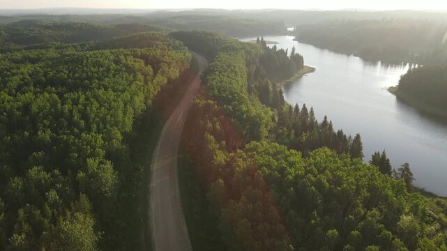 Drone flying along a northern park road beside a lake at sunrise, surrounded by forest and warm golden light in a calm wilderness setting.