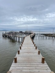 Fototapeta premium Long wooden pier stretching into calm sea under dramatic cloudy sky. Minimal coastal landscape with leading lines and peaceful atmosphere. Seaside dock and fishing platforms on horizon, travel and sol