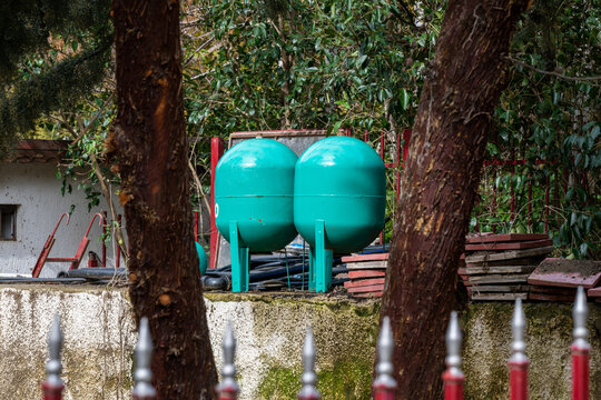 two large, teal-colored pressure tanks or expansion vessels standing behind a white concrete wall and a red metal fence, surrounded by trees and stacks of roof tiles.