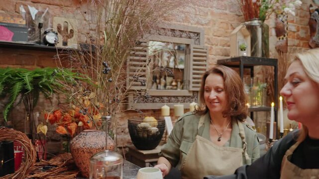 Curly white florist arranging customer order in rustic shop wearing green apron, shelves stacked with ribbons and baskets, handing single rose while chatting, scissors and twine on counter, friendly