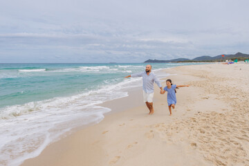 Father and daughter running happily on beach, enjoying family vacation, having fun near ocean water