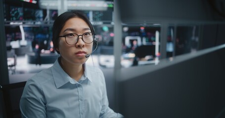 Fototapeta premium Young Asian Specialist in Crisp Blue Shirt, Focused on Real Time Analytical Dashboards. Woman Wearing Professional Headset For Instant Communication. New Generation Experts in Investment Environment.