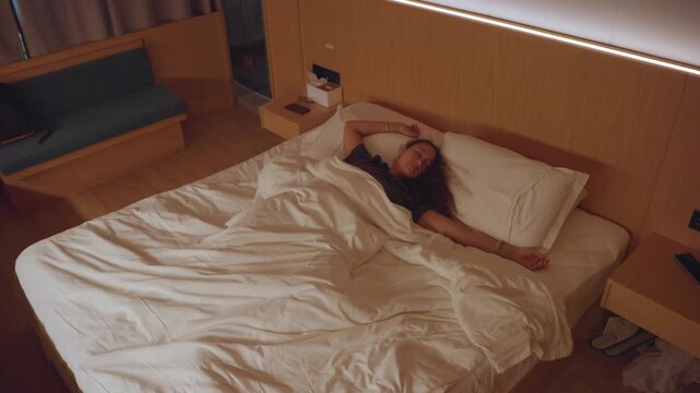 Hotel bed tired female guest waking, soft warm lighting, white duvet, overhead angle, bedside lamp, smartphone on nightstand, slow stretch, cozy boutique hotel room, intimate candid moment, solo