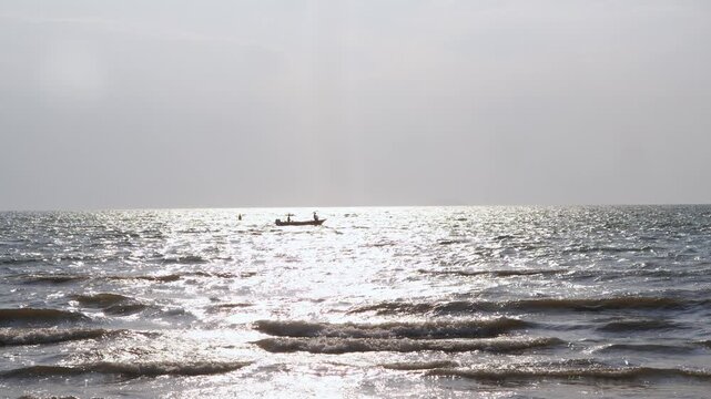 Small motorboat moves across shimmering sunlit sea surface far from shore. Gentle waves roll in foreground under bright hazy sky.