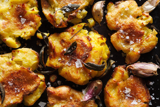 
Crispy smashed roast potatoes with garlic and sage sprinkled with salt flakes on a dark background, close up view