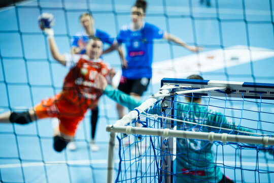 View through the handball net showing a blurred player in an orange jersey jumping to score a goal during a professional competition.