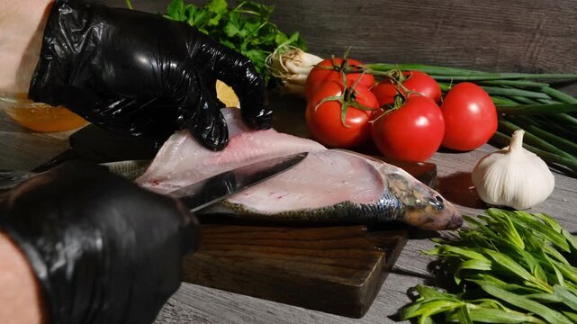 Chef filleting fish with kitchen knife on cutting board. Hands in gloves cutting fish fillet from bones, cooking fish with vegetables, spices, and lemon in seafood restaurant.
