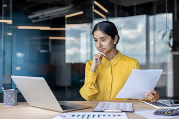Obraz premium Serious and thoughtful young Indian woman sitting at her desk. holding documents and looking at laptop screen