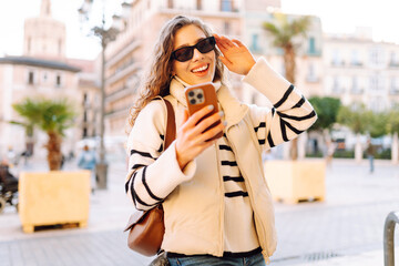 Cheerful woman wearing sunglasses and using phone strolls along sunny streets of city. Tourist...