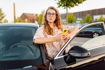 Beautiful woman wearing glasses and holding phone stands next to car on sunny day. Young woman driver stands in a parking lot holding a phone. Concept of transportation, navigation. Lifestyle. © maxbelchenko