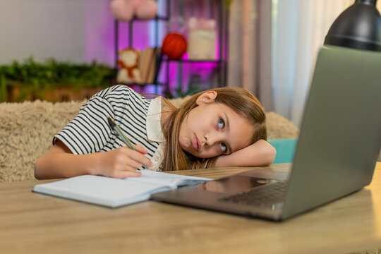 Young child girl sitting at table on home sofa, feeling tired lazy, doing homework with laptop and notebook. School kid slowly writes in exercise book struggling to stay focused during online learning