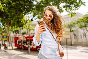 Fototapeta premium Smiling woman wearing sunglasses and using phone sits on a bench on sunny day. Young woman using her phone enjoys a walk in park and relaxes on bench. Concepts of communication, and relaxation.