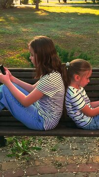 Two girls sitting back to back on a bench, Children playing on smartphones outdoors, Siblings using mobile devices in the park, Kids in striped shirts with phones, Girls focused on smartphones outside