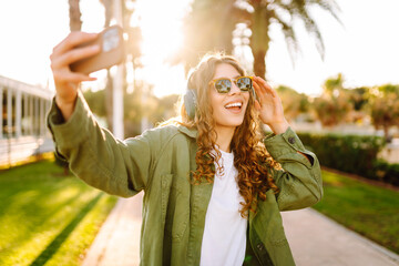 Happy woman wearing wireless headphones takes selfie on phone in a sunny park. Young woman blogs on...