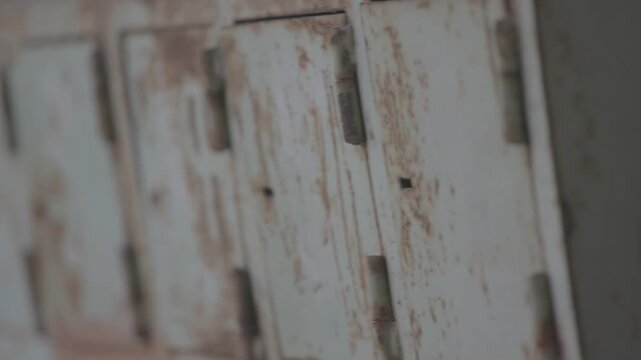 A close up view of a row of old, rusty metal mailboxes with peeling pale paint, mounted on a wall in a weathered and dilapidated outdoor urban setting.

