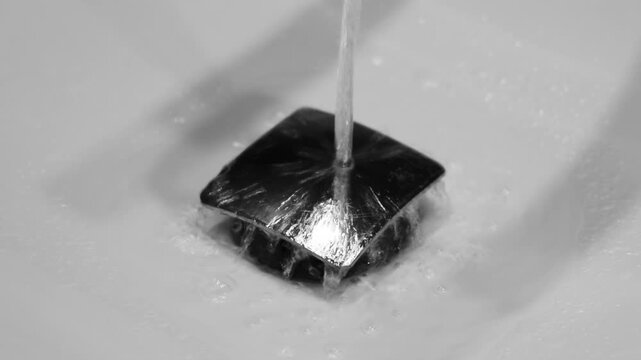 Macro close up of water stream flowing directly into square chrome sink drain. Clean white ceramic basin with modern metallic drain hole. Water disappearing down the drain