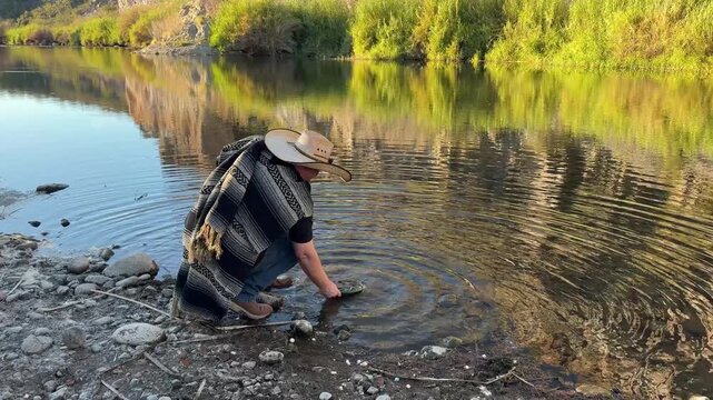 Millennial man (early 40s) wearing a poncho and cowboy hat, panning for gold in Arizona in river