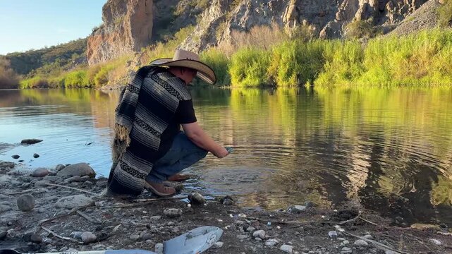 Millennial man (early 40s) wearing a poncho and cowboy hat, panning for gold in Arizona in river
