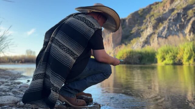 Millennial man (early 40s) wearing a poncho and cowboy hat, panning for gold in Arizona in river