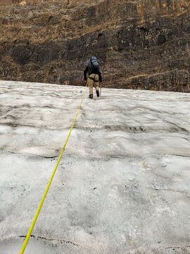 A man wearing a backpack and tethered to a rope, climbs a glacier in the rocky mountains with an icepick during summer.