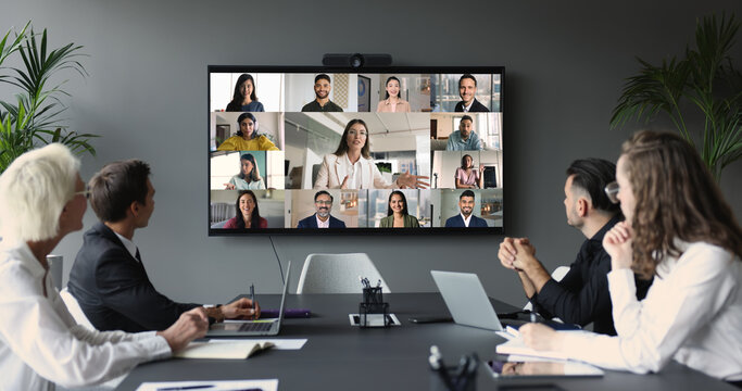 Group of businesspeople gather at conference table collaborating with multiple participants from diverse locations displayed on TV screen, take part in corporate discussion. Business virtual meeting
