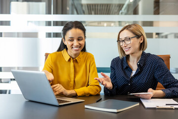 Two smiling multiethnic businesswomen collaborating on a project, viewing a laptop screen and...