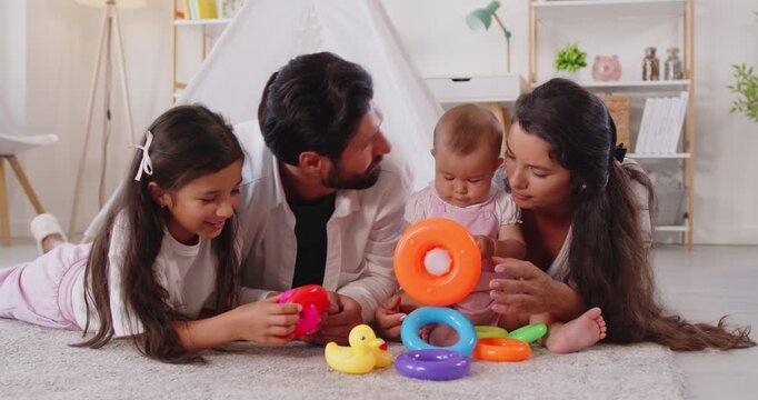 Family with children play educational game with toys at home. Sister and toddler baby explore colorful stacking rings pyramid on floor with parents. Learning, boosting development and leisure. 