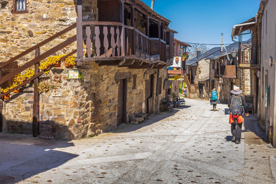 Pilgrims walk through the narrow stone streets of El Acebo de San Miguel featuring traditional wooden balconies and flower-lined stairs, historic architecture on the Camino de Santiago, Spain