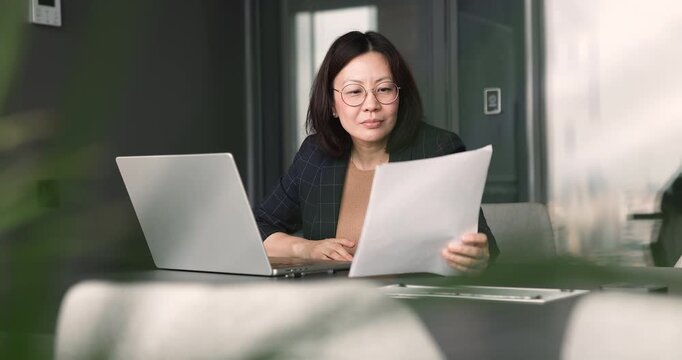 Focused middle-aged 50s Asian businesswoman typing on laptop and reviewing printed document, working on financial report, preparing business documentation, comparing data, updating records in office