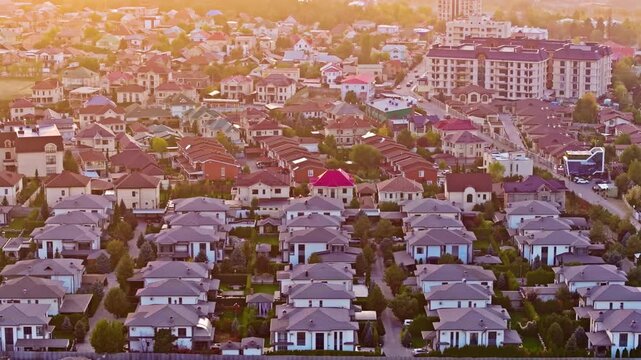 Aerial drone shot trucking right over Sary Ozon cottage settlement in Kyrgyzstan at sunset, showing dense detached houses, tiled roofs, inner roads and mid-rise buildings.