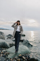 Woman in coat and hat stands on rocky shore near water under cloudy sky. Casual fashion, autumn style, and outdoor mood with calm sea and natural landscape background. © SHOTPRIME STUDIO