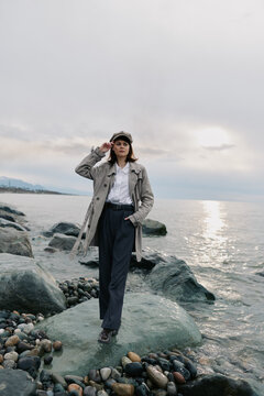 Woman in coat and hat stands on rocky shore near water under cloudy sky. Casual fashion, autumn style, and outdoor mood with calm sea and natural landscape background.