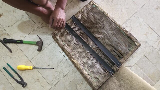 Top view of a handyman repairing a vintage wooden chair seat by attaching black elastic webbing straps with a staple gun during furniture restoration work on a tiled floor