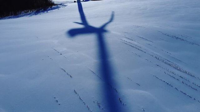 Aerial view moving away from the shadow of a wind turbine spinning at full speed, stretching lengthwise across a snow-covered agricultural field. Saint-Leandre, Quebec, Canada.
